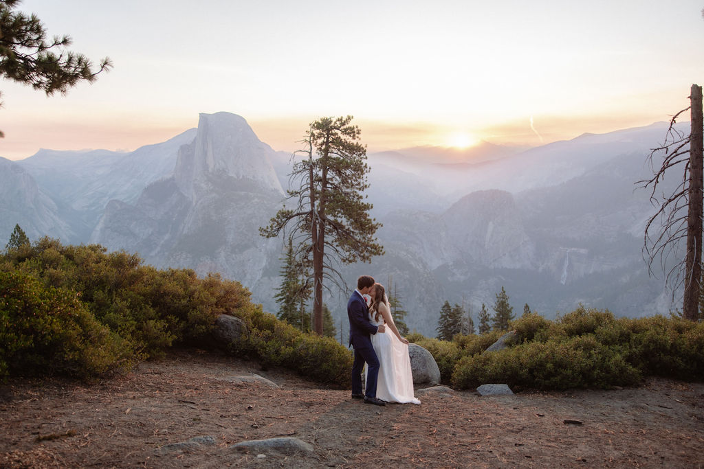A bride in a white dress stands beside a groom in a suit on a rocky ledge, with mountains and trees in the background for a wedding at glacier point