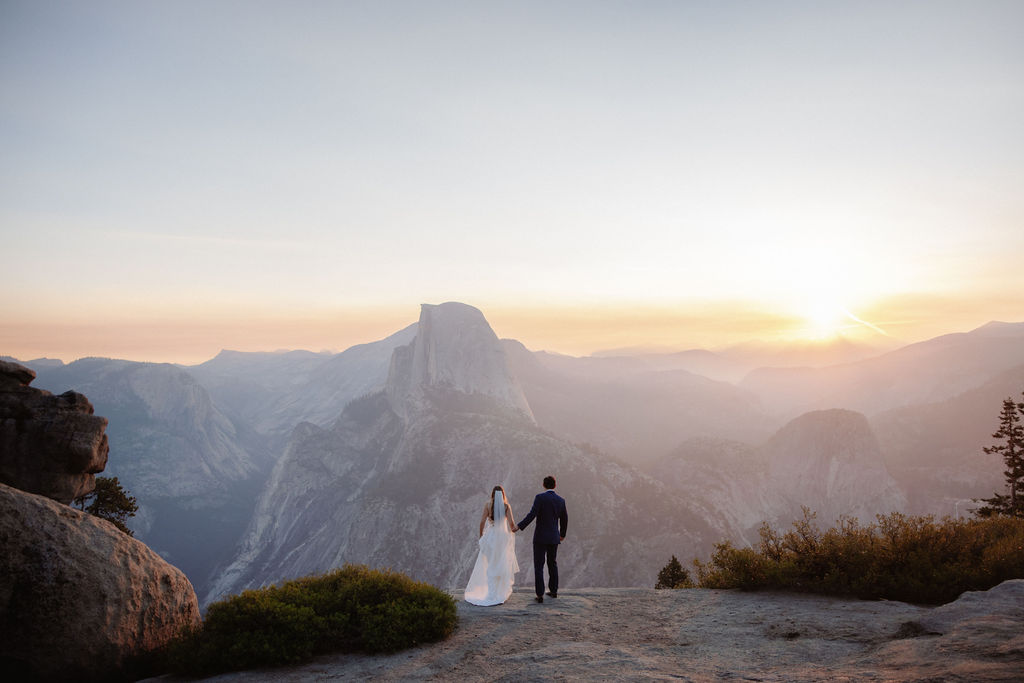 A bride in a white dress stands beside a groom in a suit on a rocky ledge, with mountains and trees in the background for a wedding at glacier point