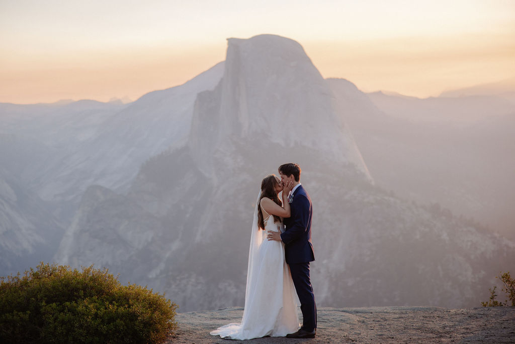 A bride and groom stand holding hands on a rocky ledge with mountains and a large rock formation in the background at sunset.