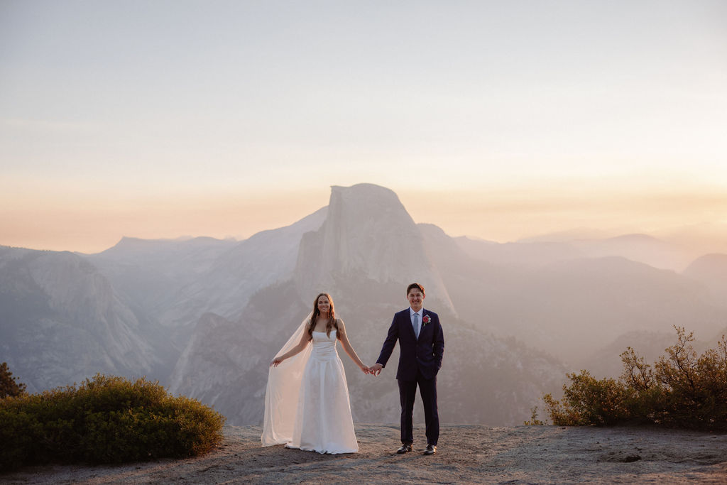 A bride and groom stand holding hands on a rocky ledge with mountains and a large rock formation in the background at sunset.