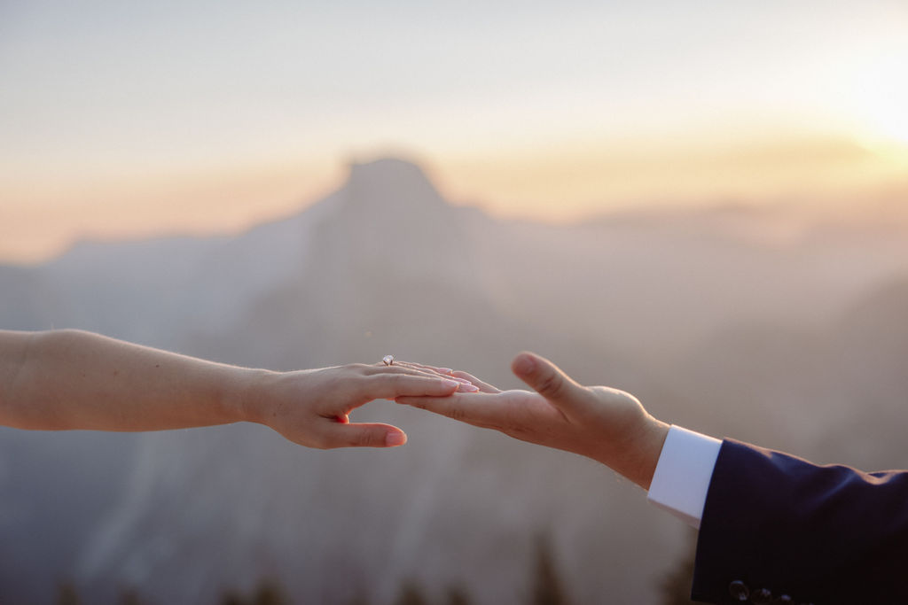 A bride in a white dress stands beside a groom in a suit on a rocky ledge, with mountains and trees in the background for a wedding at glacier point