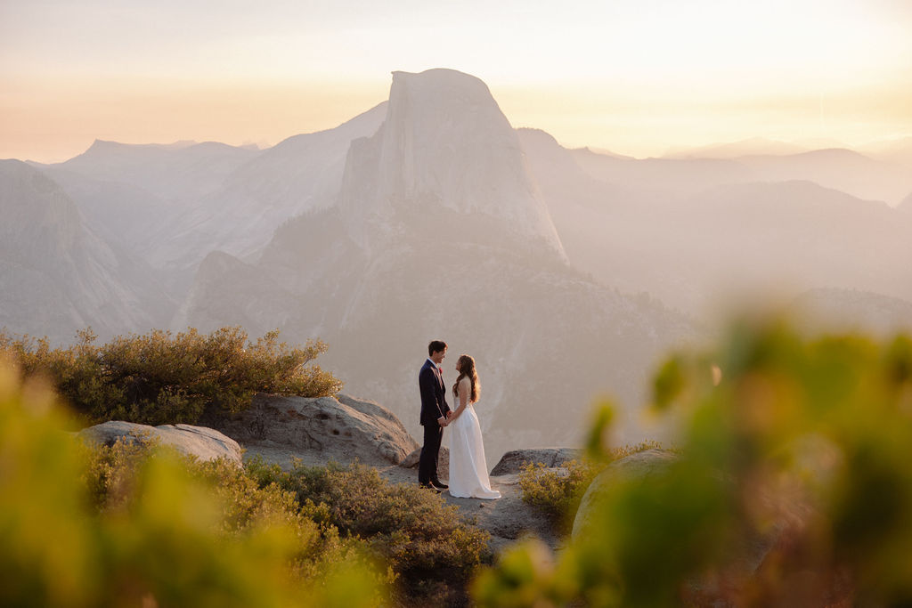 A bride in a white dress stands beside a groom in a suit on a rocky ledge, with mountains and trees in the background for a wedding at glacier point