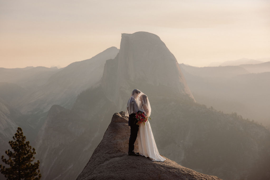 A bride and groom share a kiss on a rocky cliff with mountains and Half Dome in the background under a hazy sky for a glacier point wedding