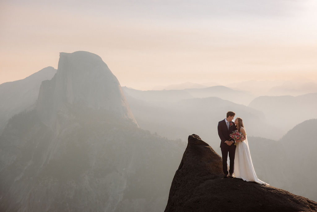A bride and groom share a kiss on a rocky cliff with mountains and Half Dome in the background under a hazy sky for a glacier point wedding
