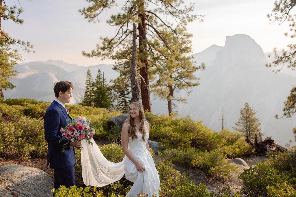 A bride and groom stand hand in hand outdoors, facing a scenic mountain landscape with trees and mist in the background.