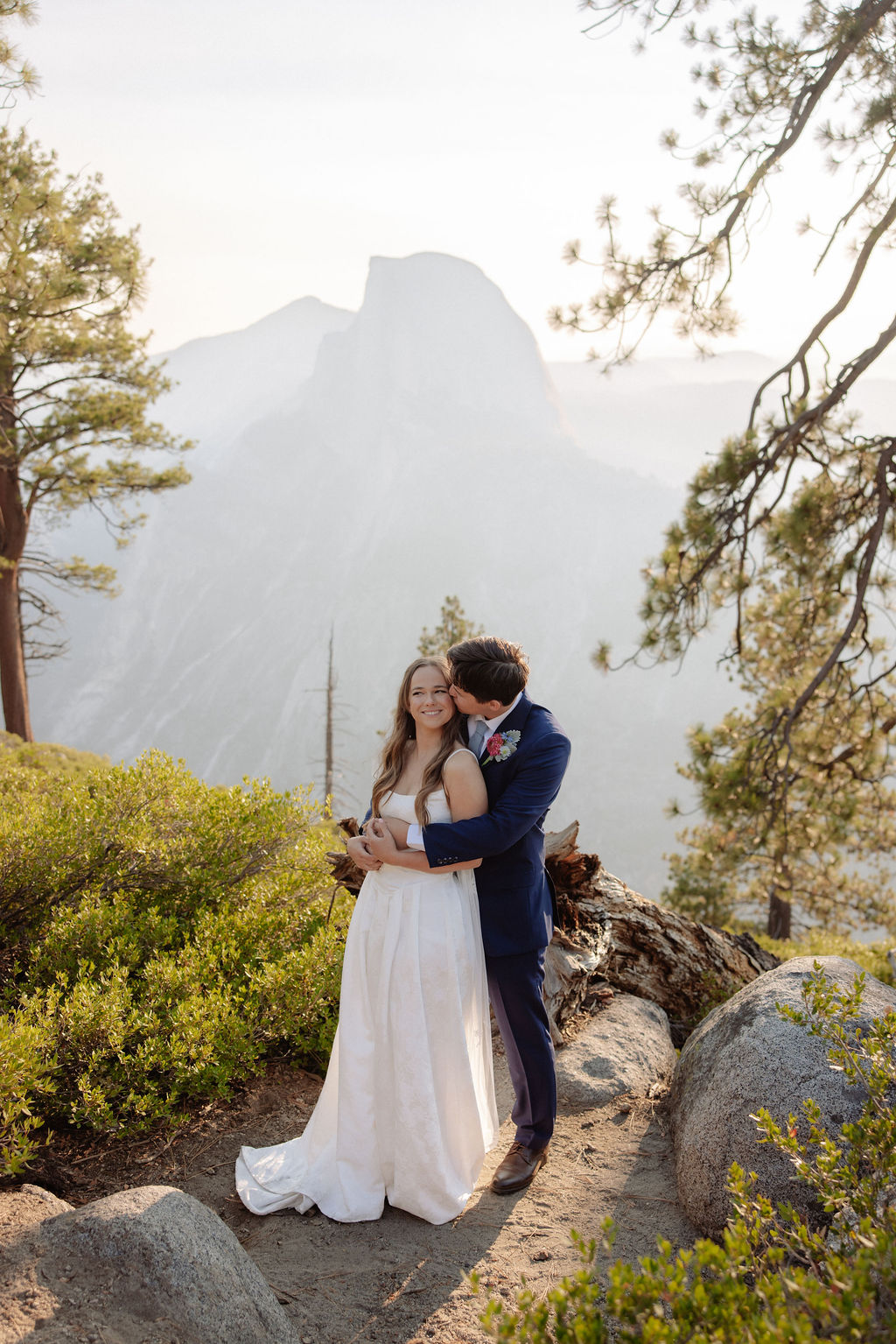 A bride and groom stand hand in hand outdoors, facing a scenic mountain landscape with trees and mist in the background.