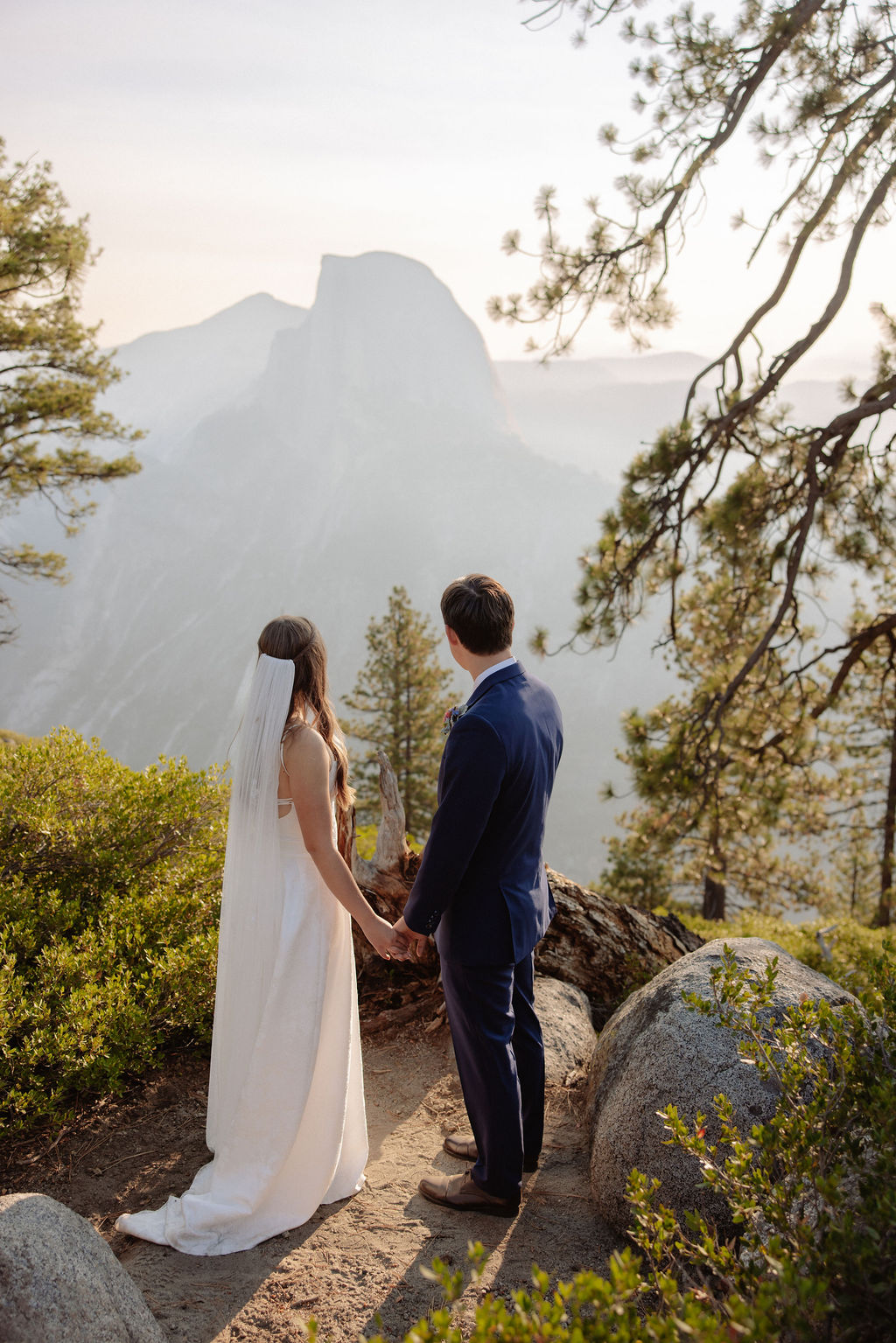 A bride and groom stand hand in hand outdoors, facing a scenic mountain landscape with trees and mist in the background.