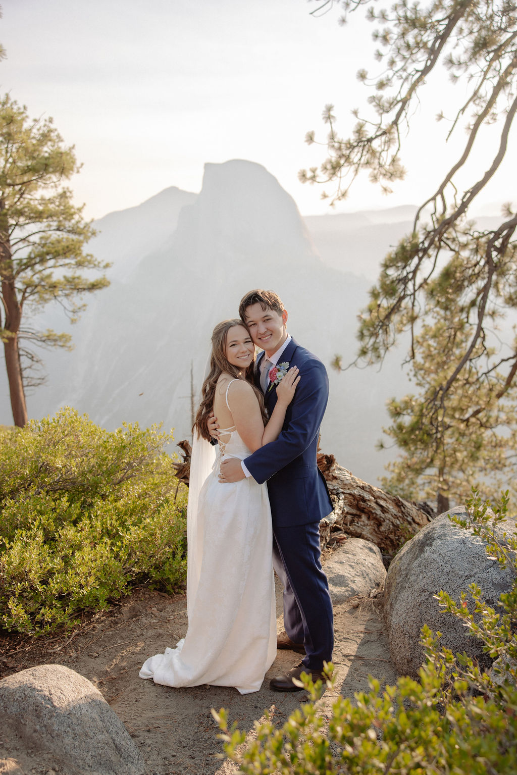 A bride and groom stand hand in hand outdoors, facing a scenic mountain landscape with trees and mist in the background.