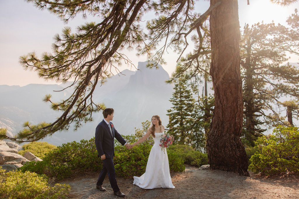A bride and groom stand hand in hand outdoors, facing a scenic mountain landscape with trees and mist in the background.