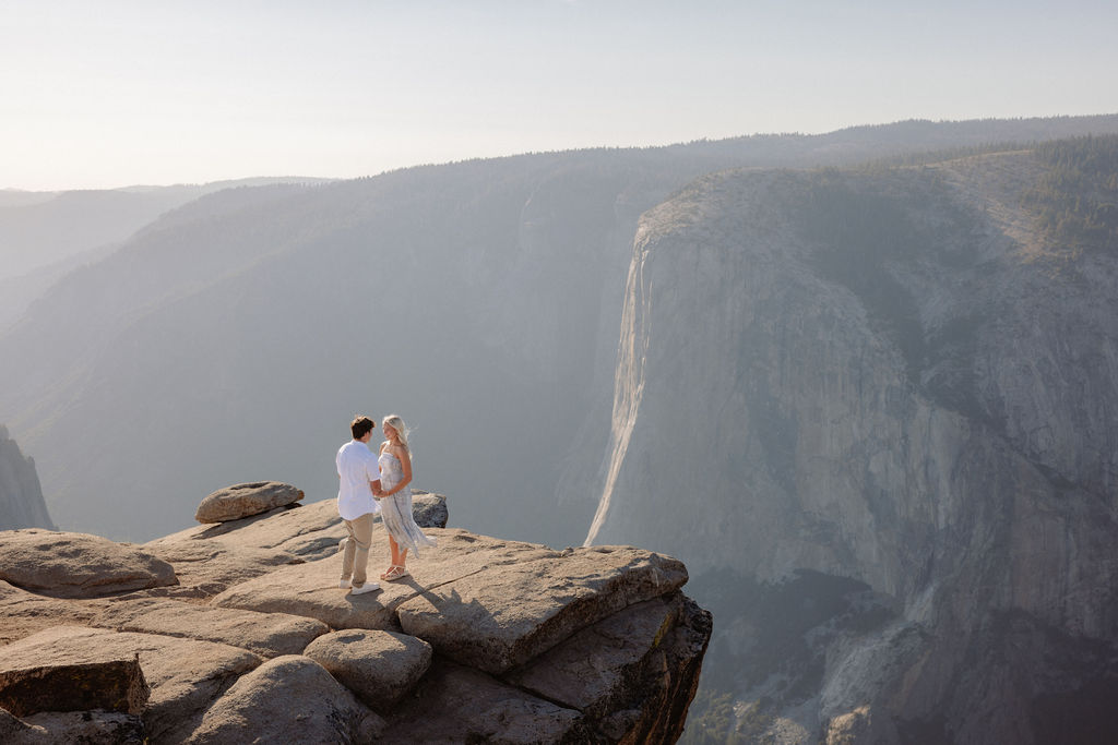 A person kneels and proposes to another person dressed in white on a rocky cliff with mountains in the background for a Yosemite National Park Proposal