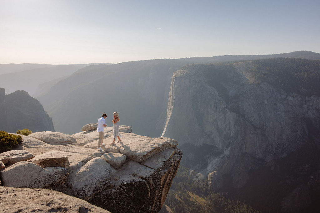 A person kneels and proposes to another person dressed in white on a rocky cliff with mountains in the background for a Yosemite National Park Proposal