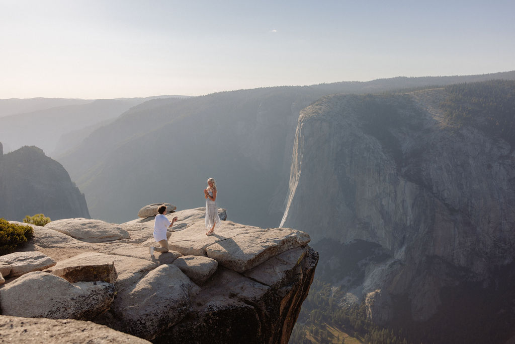 A person kneels and proposes to another person dressed in white on a rocky cliff with mountains in the background for a Yosemite National Park Proposal