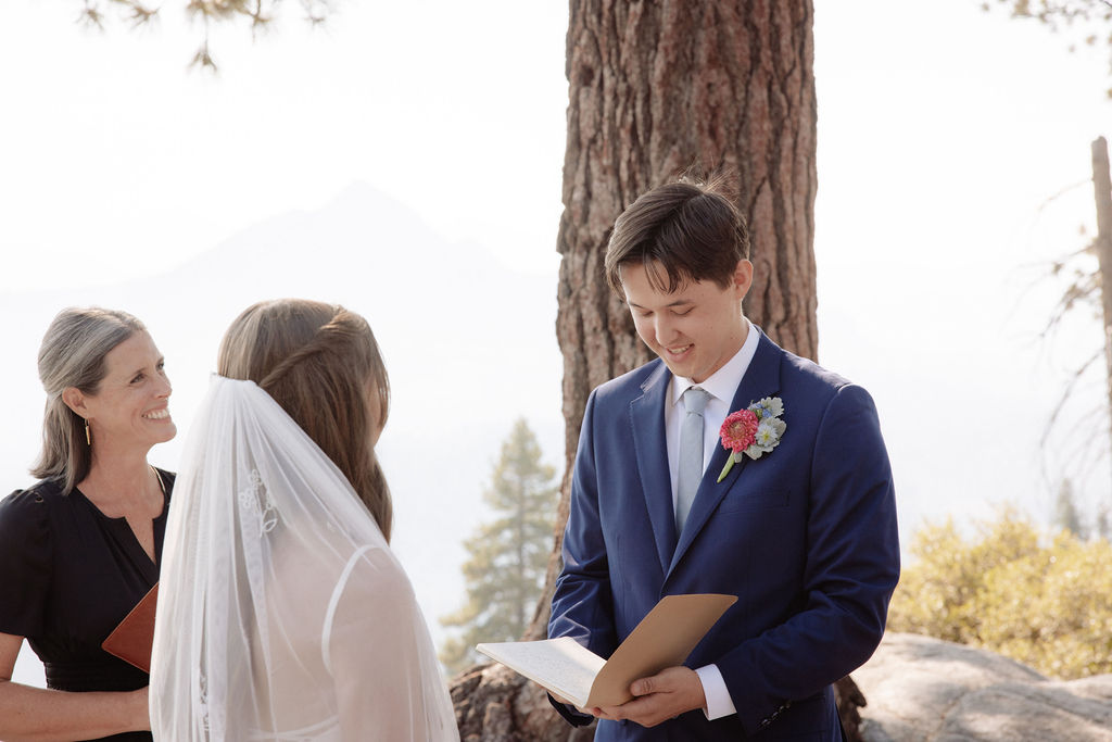 A couple stands with an officiant on a rocky overlook, surrounded by trees and mountains, while guests watch the outdoor wedding ceremony.