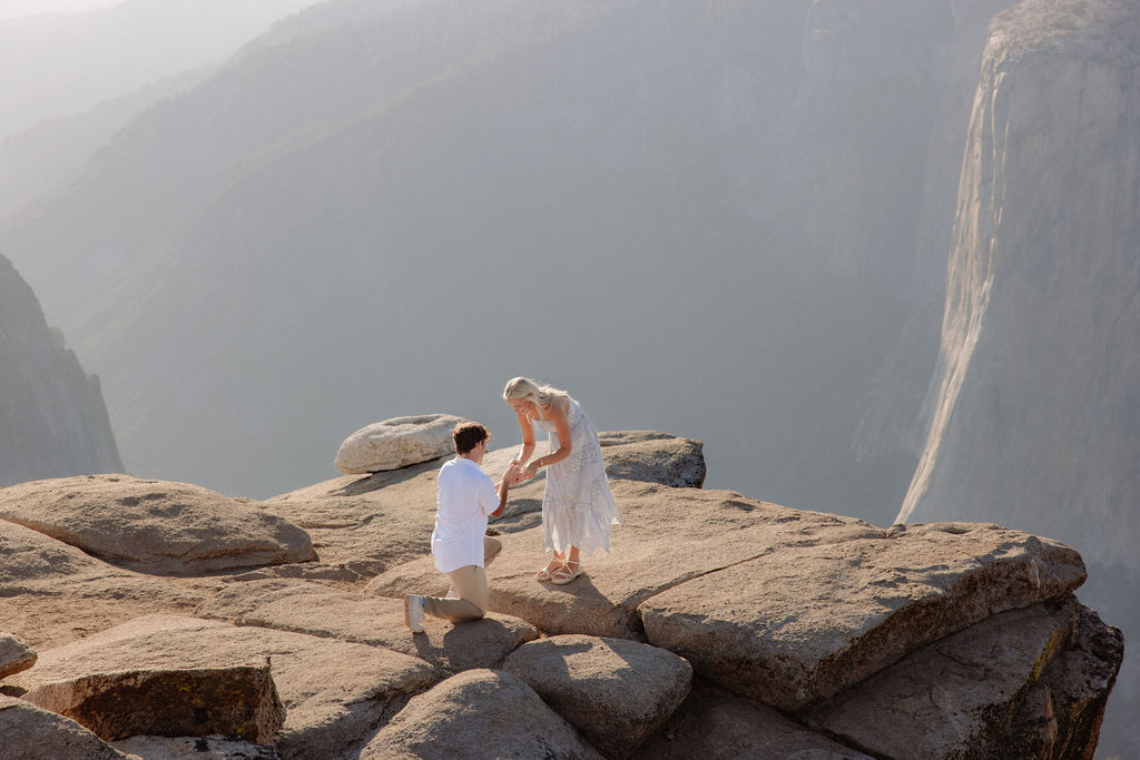 A person kneels and proposes to another person dressed in white on a rocky cliff with mountains in the background for a Yosemite National Park Proposal