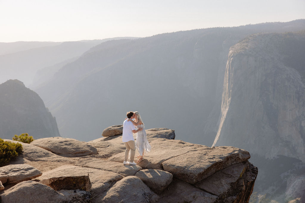 A person kneels and proposes to another person dressed in white on a rocky cliff with mountains in the background for a Yosemite National Park Proposal