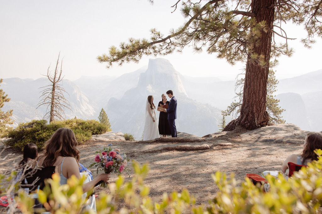 A couple stands with an officiant on a rocky overlook, surrounded by trees and mountains, while guests watch the outdoor wedding ceremony.