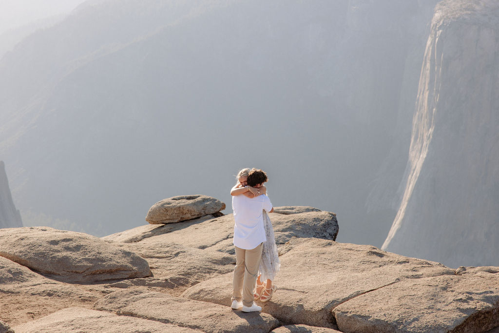 A person kneels and proposes to another person dressed in white on a rocky cliff with mountains in the background for a Yosemite National Park Proposal