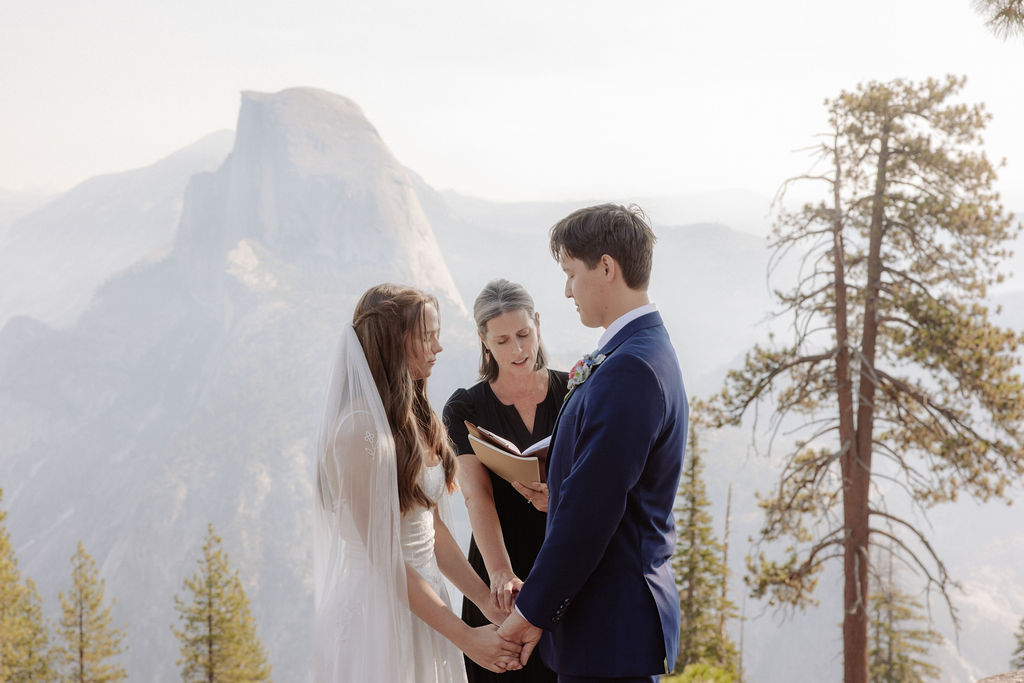 A couple stands with an officiant on a rocky overlook, surrounded by trees and mountains, while guests watch the outdoor wedding ceremony.