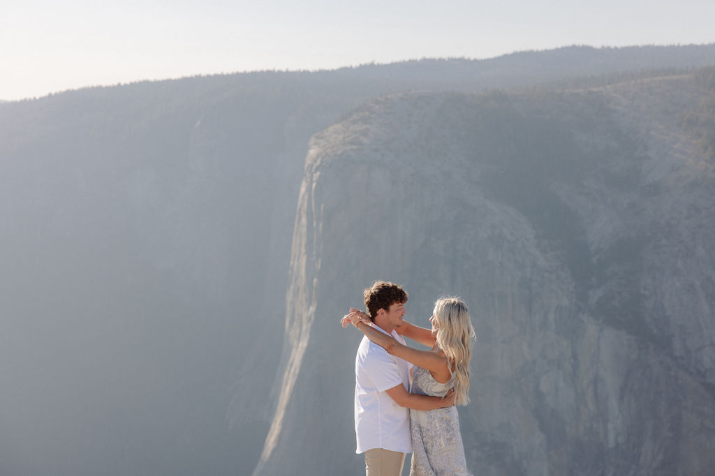 Two people sit closely together on the edge of a rocky cliff with a vast mountainous landscape in the background for a Yosemite national park proposal