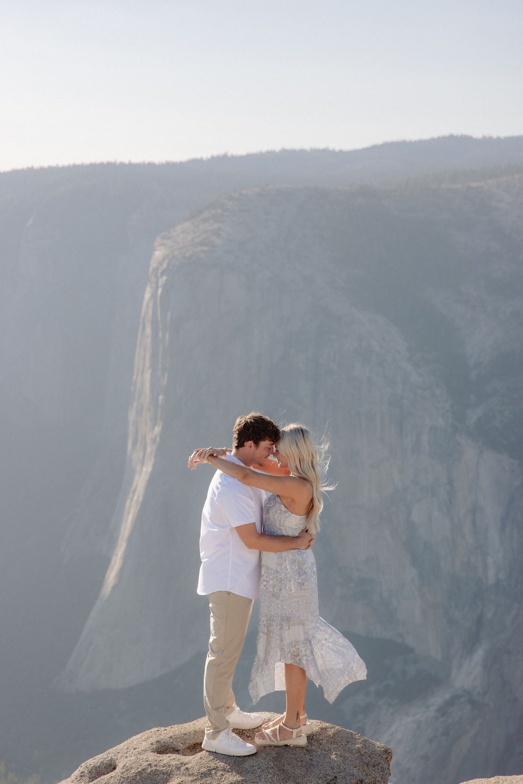 Two people sit closely together on the edge of a rocky cliff with a vast mountainous landscape in the background for a Yosemite national park proposal