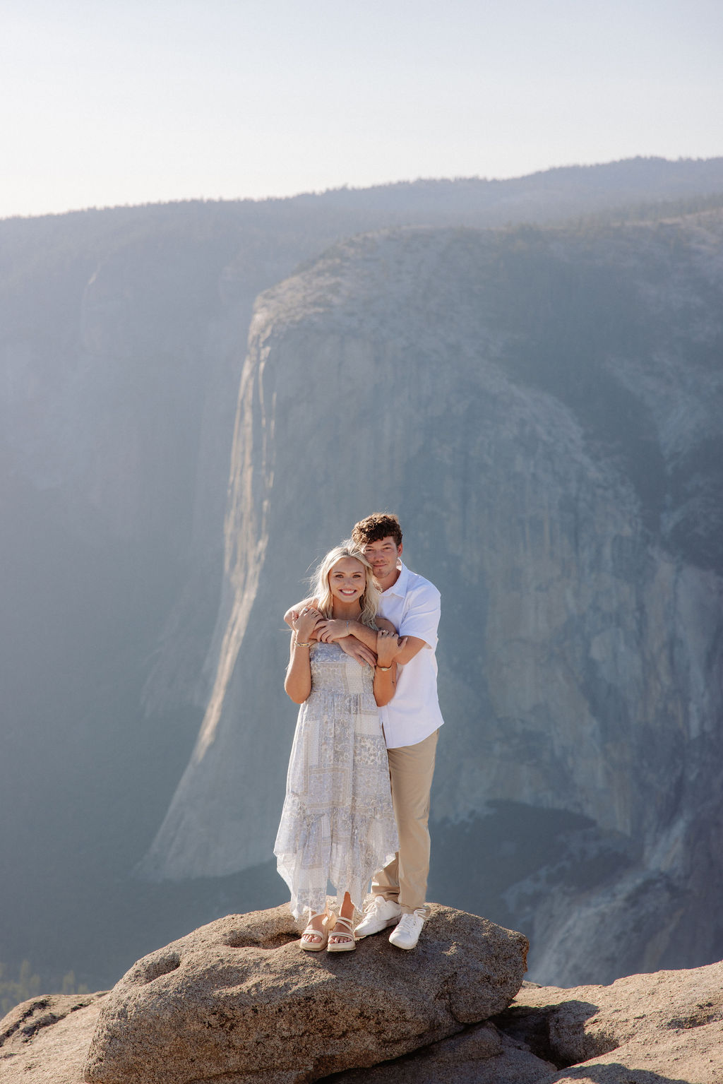 Two people sit closely together on the edge of a rocky cliff with a vast mountainous landscape in the background for a Yosemite national park proposal