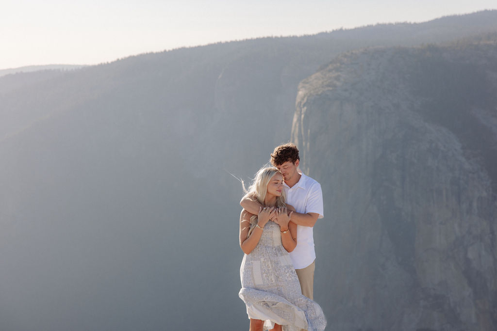 Two people sit closely together on the edge of a rocky cliff with a vast mountainous landscape in the background for a Yosemite national park proposal