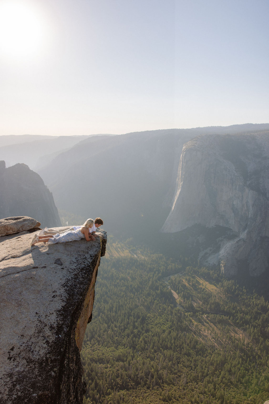 Two people sit closely together on the edge of a rocky cliff with a vast mountainous landscape in the background for a Yosemite national park proposal
