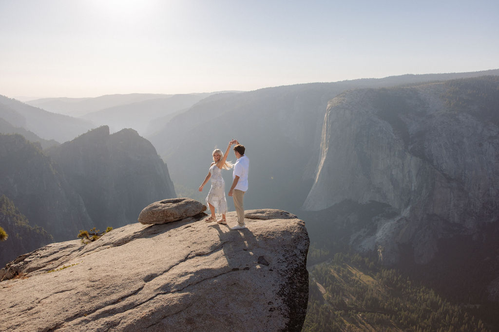 Two people sit closely together on the edge of a rocky cliff with a vast mountainous landscape in the background for a Yosemite national park proposal