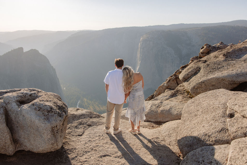 Two people sit closely together on the edge of a rocky cliff with a vast mountainous landscape in the background for a Yosemite national park proposal