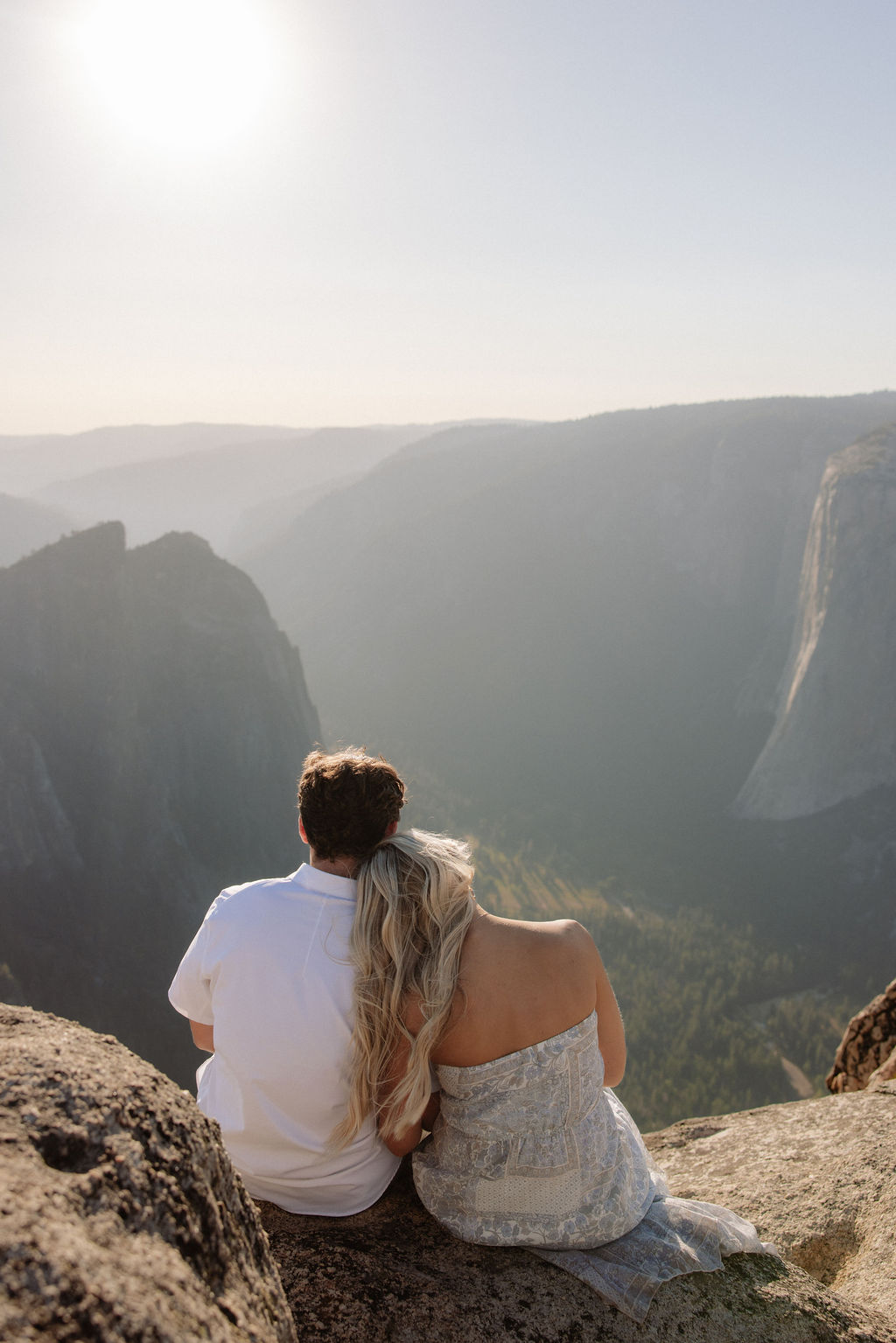 Two people sit closely together on the edge of a rocky cliff with a vast mountainous landscape in the background for a Yosemite national park proposal