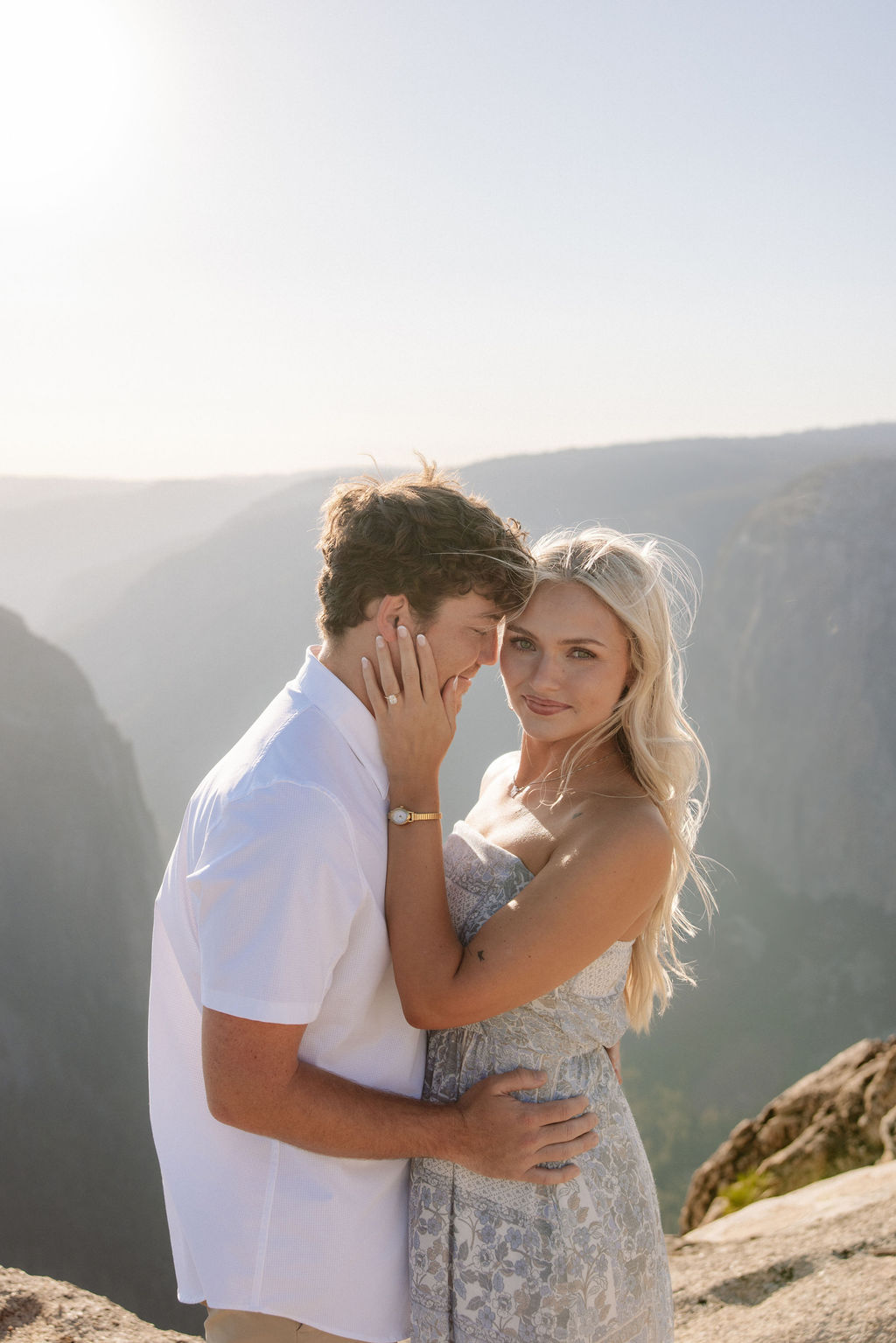 Two people sit closely together on the edge of a rocky cliff with a vast mountainous landscape in the background for a Yosemite national park proposal