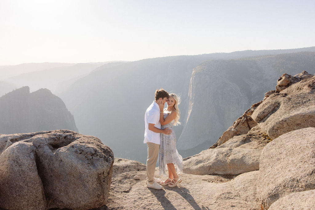 A couple stands close together on a rocky cliff with mountains and a hazy sky in the background.