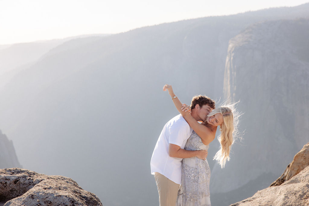 A couple stands close together on a rocky cliff with mountains and a hazy sky in the background.