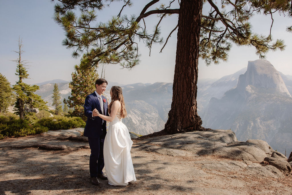 A bride and groom stand together on a rocky overlook with tall pine trees and mountains in the background.