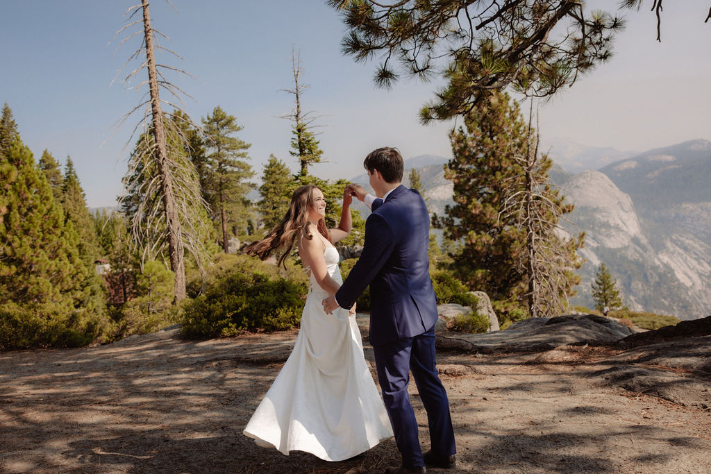 A bride and groom stand together on a rocky overlook with tall pine trees and mountains in the background.