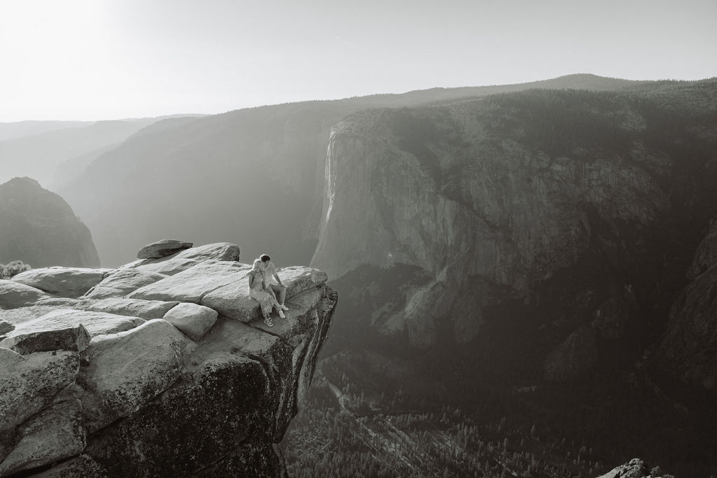 A couple stands close together on a rocky cliff with mountains and a hazy sky in the background.