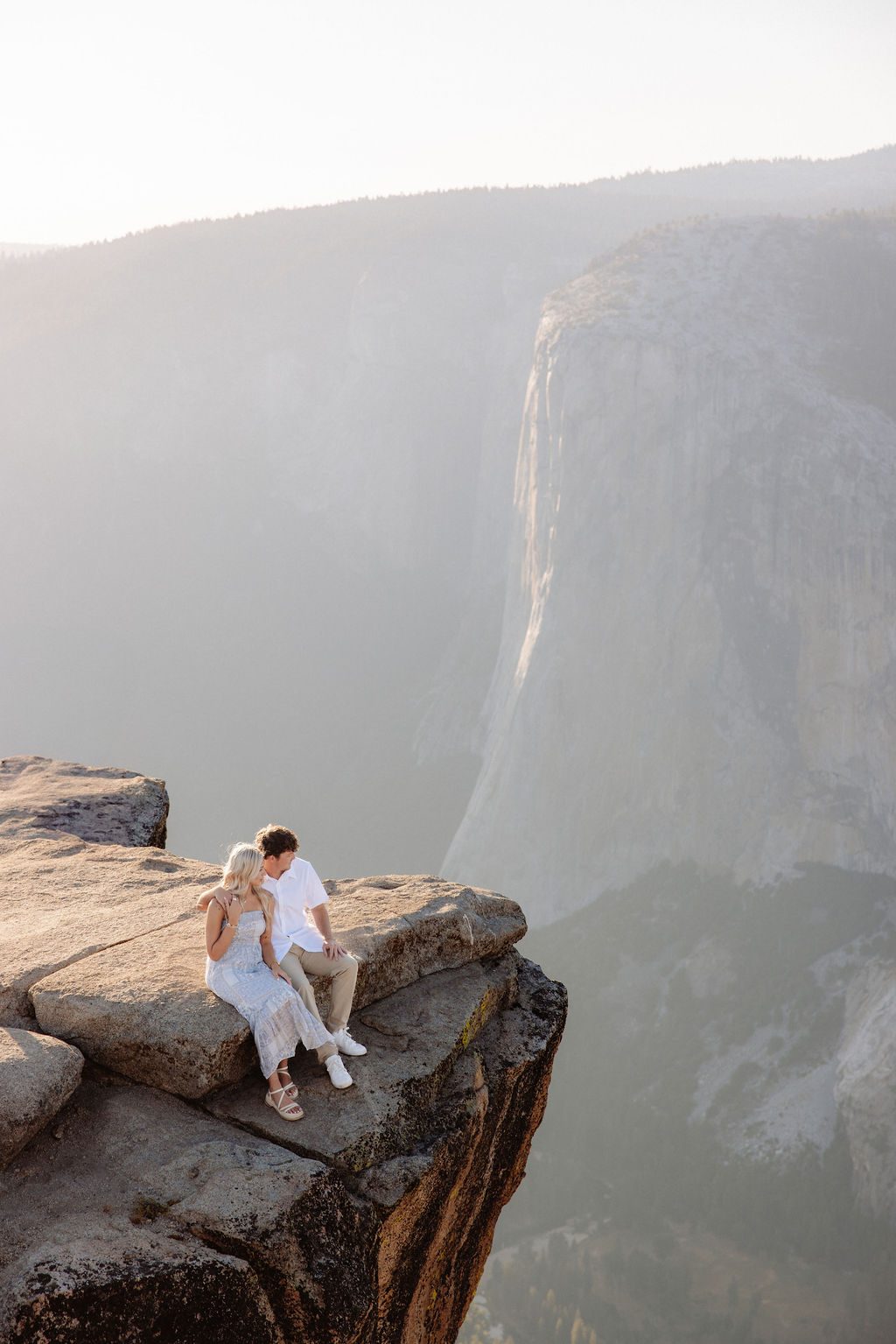 Two people sit closely together on the edge of a rocky cliff with a vast mountainous landscape in the background for a Yosemite national park proposal