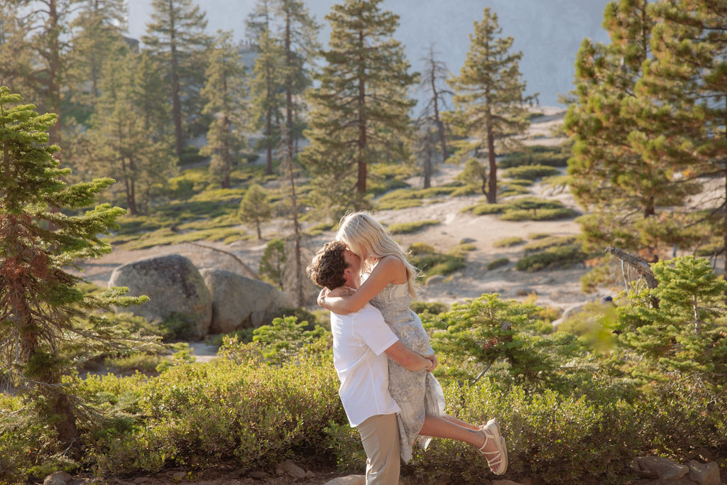 A man lifts a woman in an embrace outdoors, surrounded by trees and greenery in a sunlit, forested area.