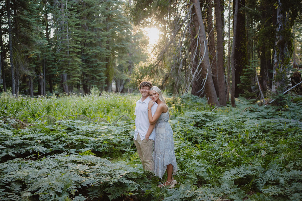 A couple stands together in a sunlit forest clearing, surrounded by tall trees and green foliage.