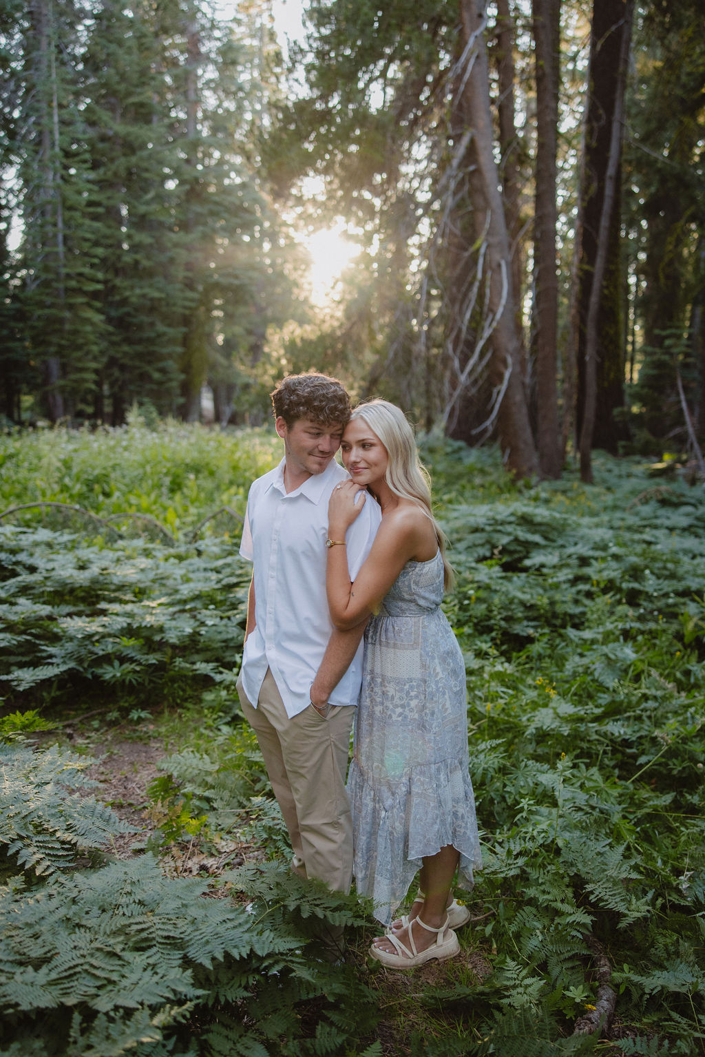 A couple stands together in a sunlit forest clearing, surrounded by tall trees and green foliage.