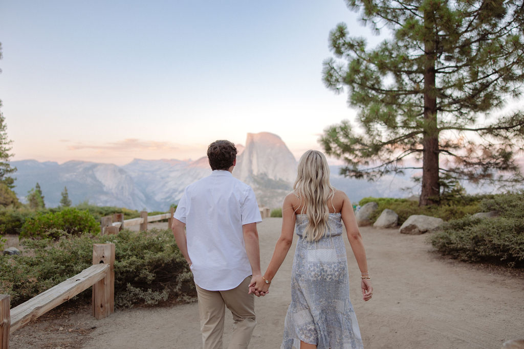 A man and woman are running playfully near a large tree with mountains and a rock formation in the background.