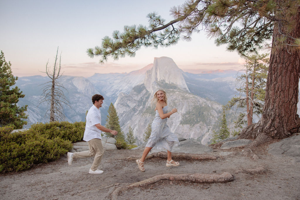 A man and woman are running playfully near a large tree with mountains and a rock formation in the background.