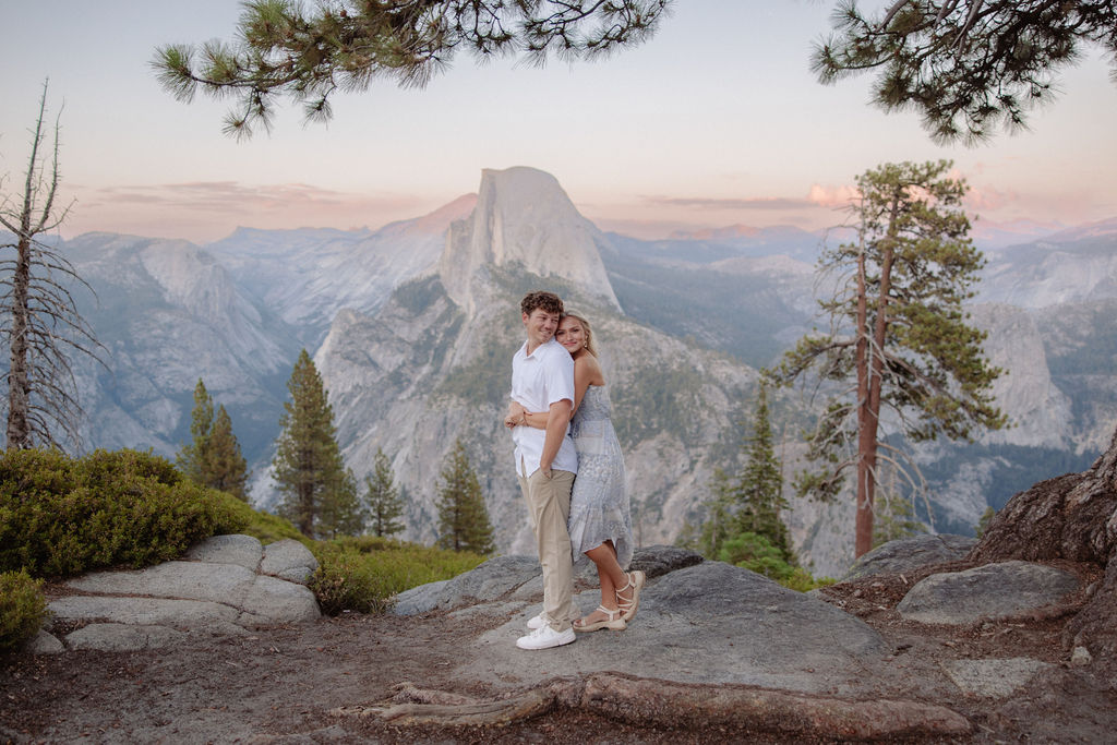 A man and woman are running playfully near a large tree with mountains and a rock formation in the background.