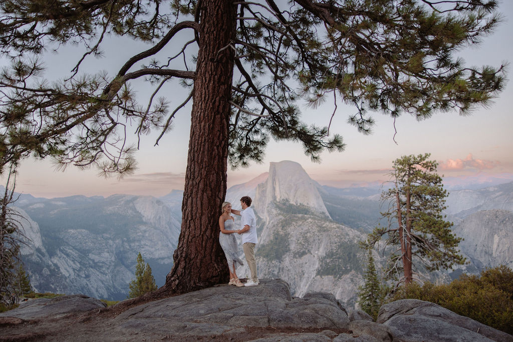 A couple stands under a large pine tree on a rocky overlook with mountains and Half Dome visible in the background.