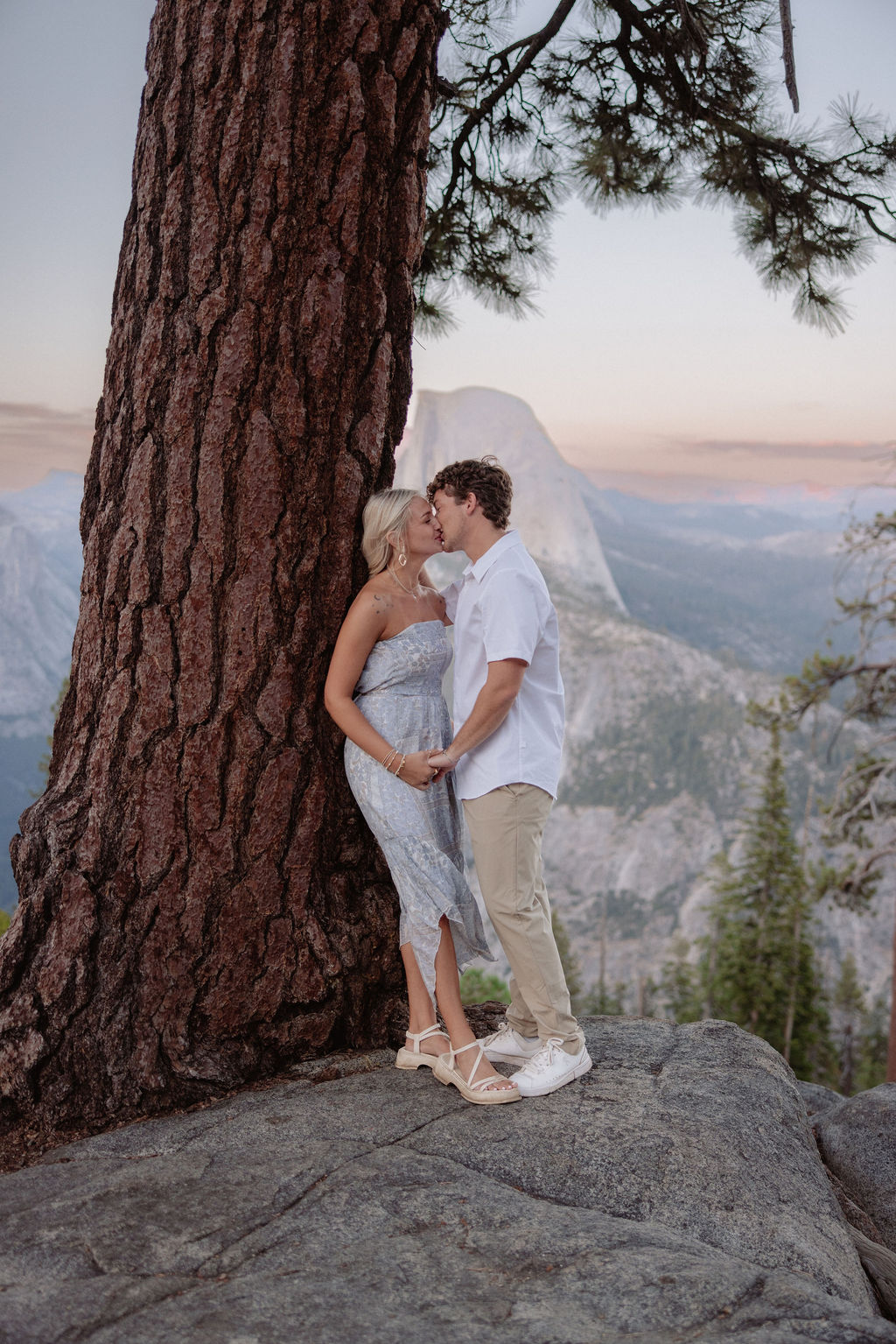 A couple stands under a large pine tree on a rocky overlook with mountains and Half Dome visible in the background.