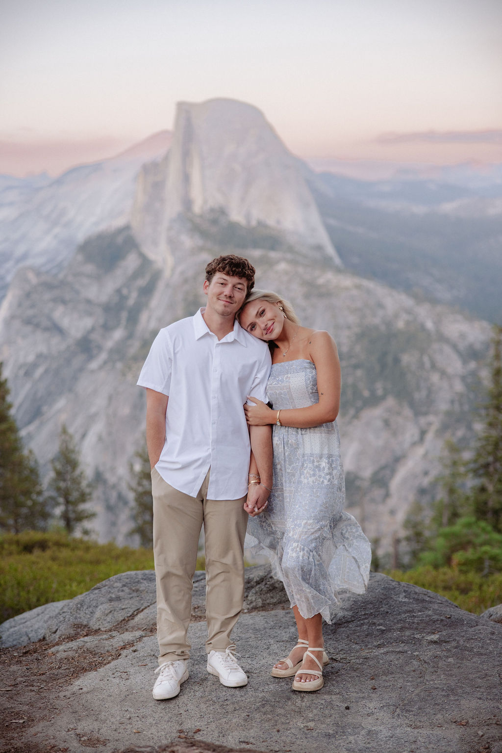 A couple stands under a large pine tree on a rocky overlook with mountains and Half Dome visible in the background for a yosemite national park proposal