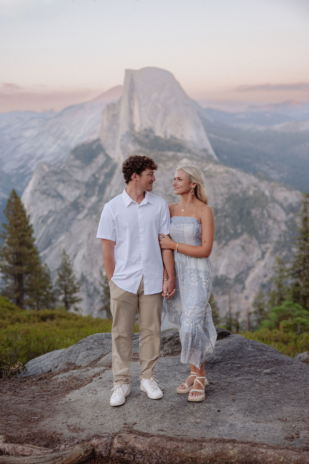 A couple stands under a large pine tree on a rocky overlook with mountains and Half Dome visible in the background for a yosemite national park proposal