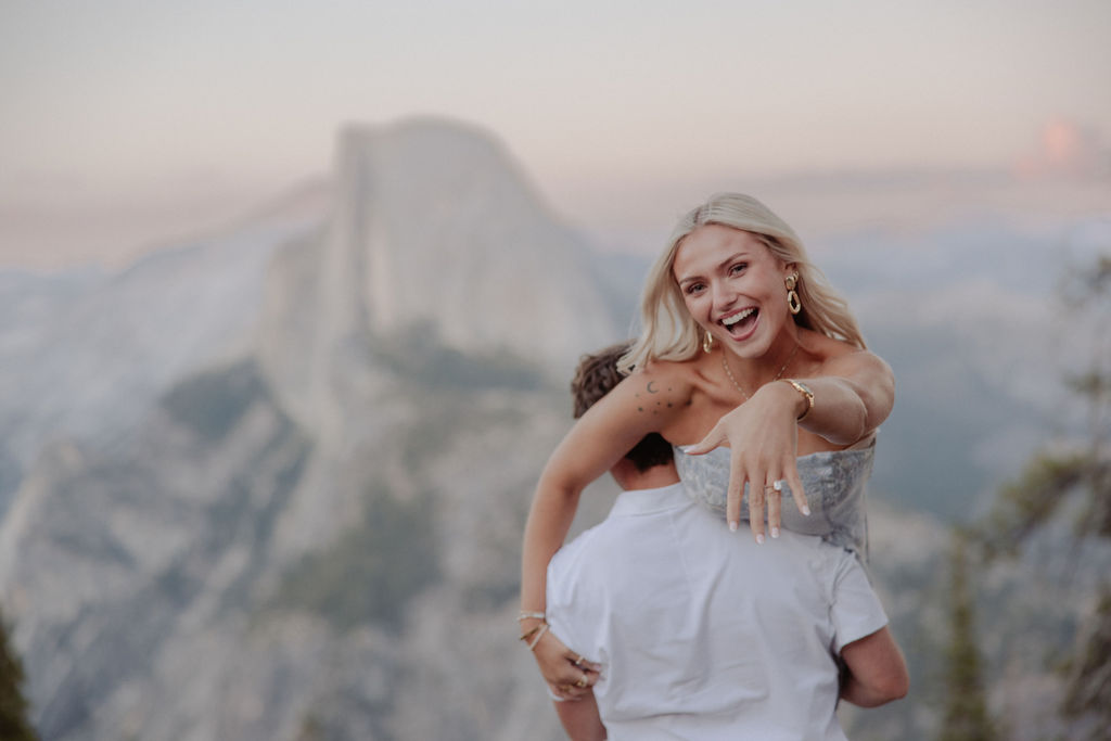 A woman shows off an engagement ring while being carried by a man, with a mountain landscape in the background.