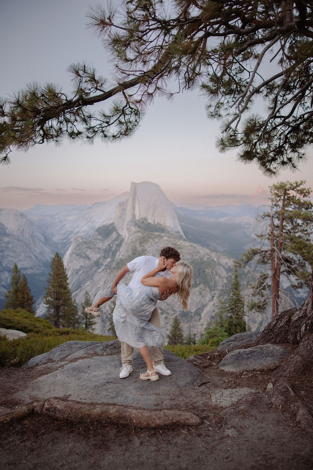 A couple stands under a large pine tree on a rocky overlook with mountains and Half Dome visible in the background for a yosemite national park proposal