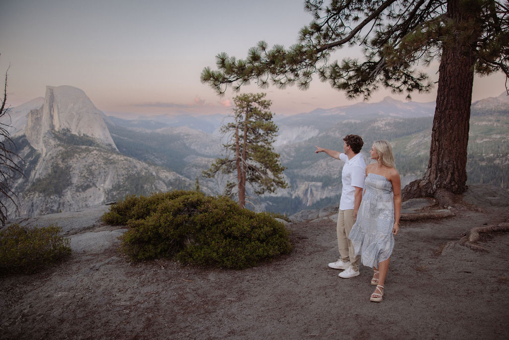 A couple stands under a large pine tree on a rocky overlook with mountains and Half Dome visible in the background for a yosemite national park proposal